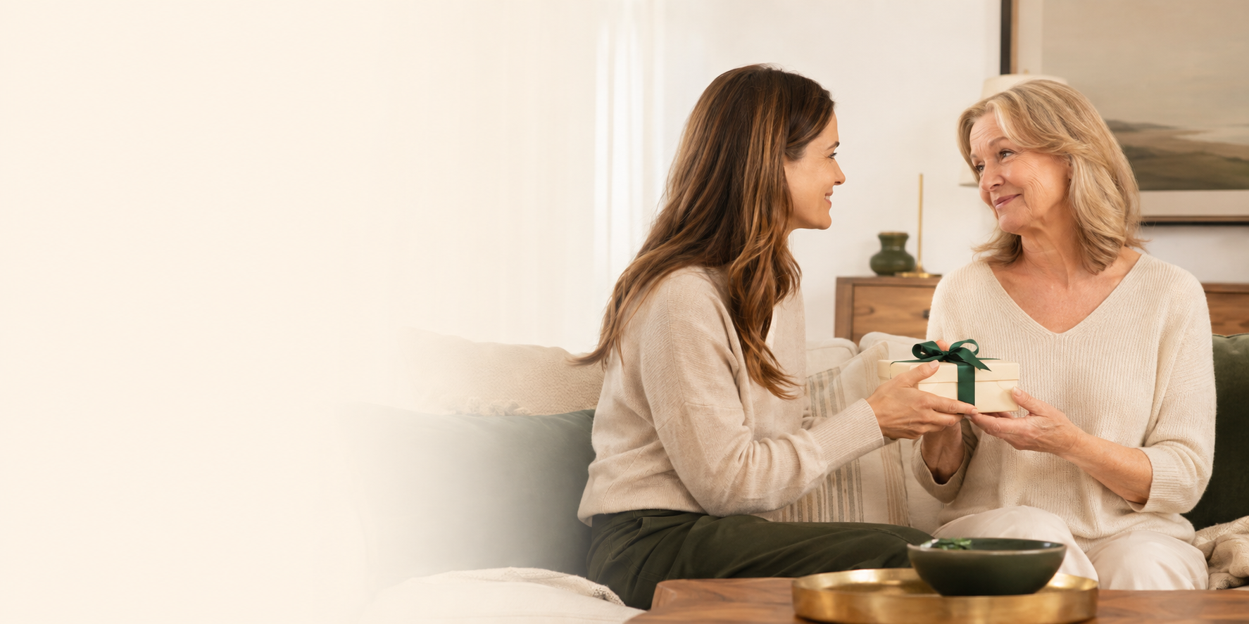 A daughter hands her mother a ribboned keepsake gift box on a sofa in warm daylight